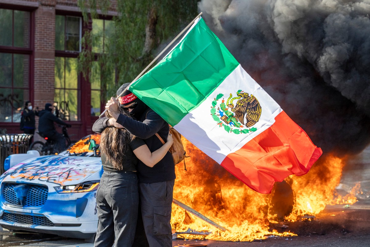 Una pareja con una bandera se abraza mientras un vehículo de Waymo arde, en medio de enfrentamientos entre manifestantes y las fuerzas del orden en las calles alrededor del edificio federal, durante una protesta tras las operaciones de inmigración federal en Los Ángeles, California, el 8 de junio de 2025 Foto: AFP
